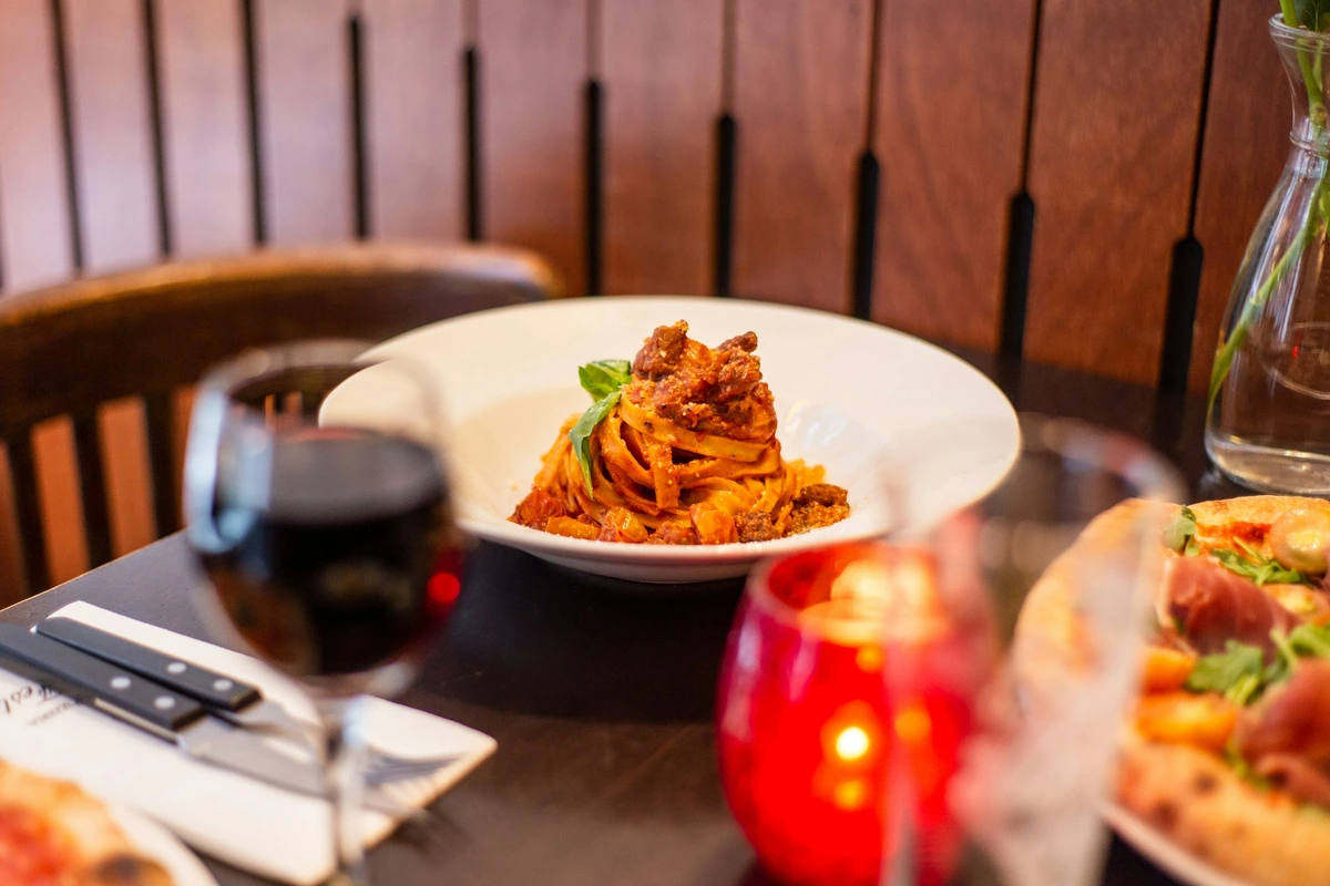 Massa ao ragù em prato branco, com taça de vinho tinto desfocada em primeiro plano