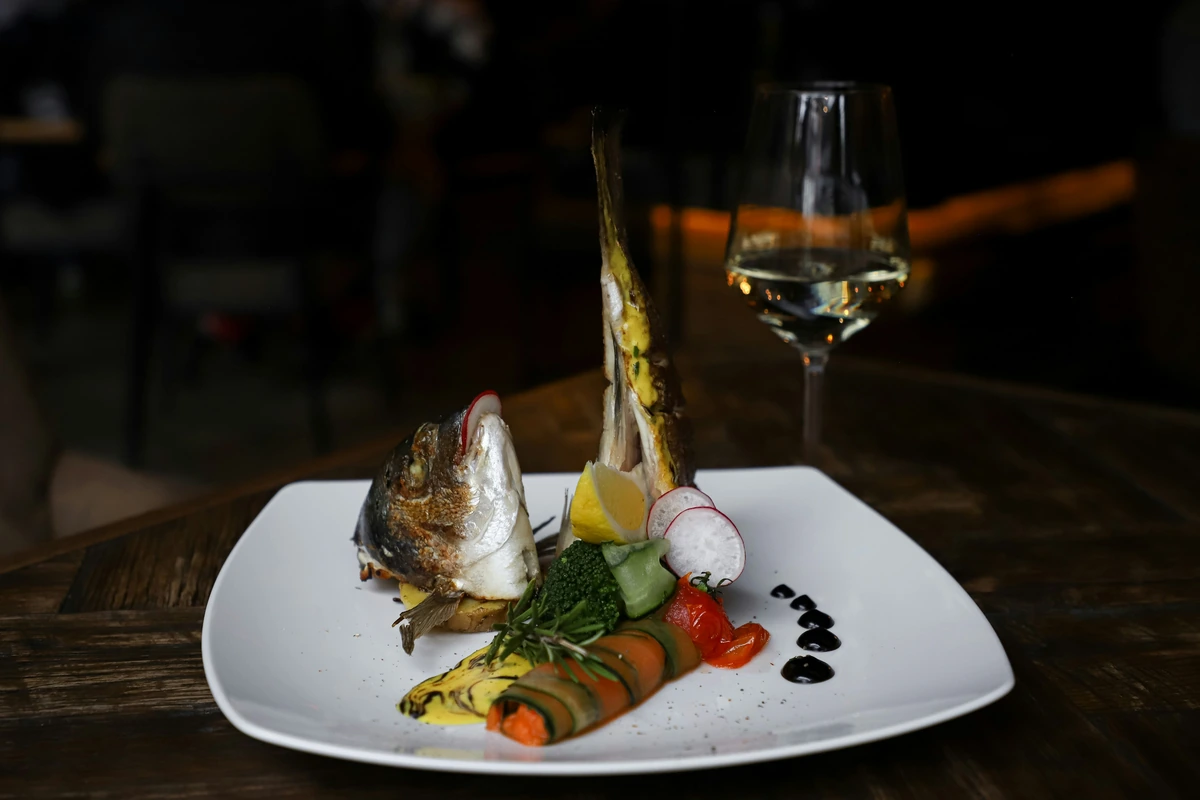 Preparação de peixe com legumes e guarnições acompanhada de taça de vinho branco.