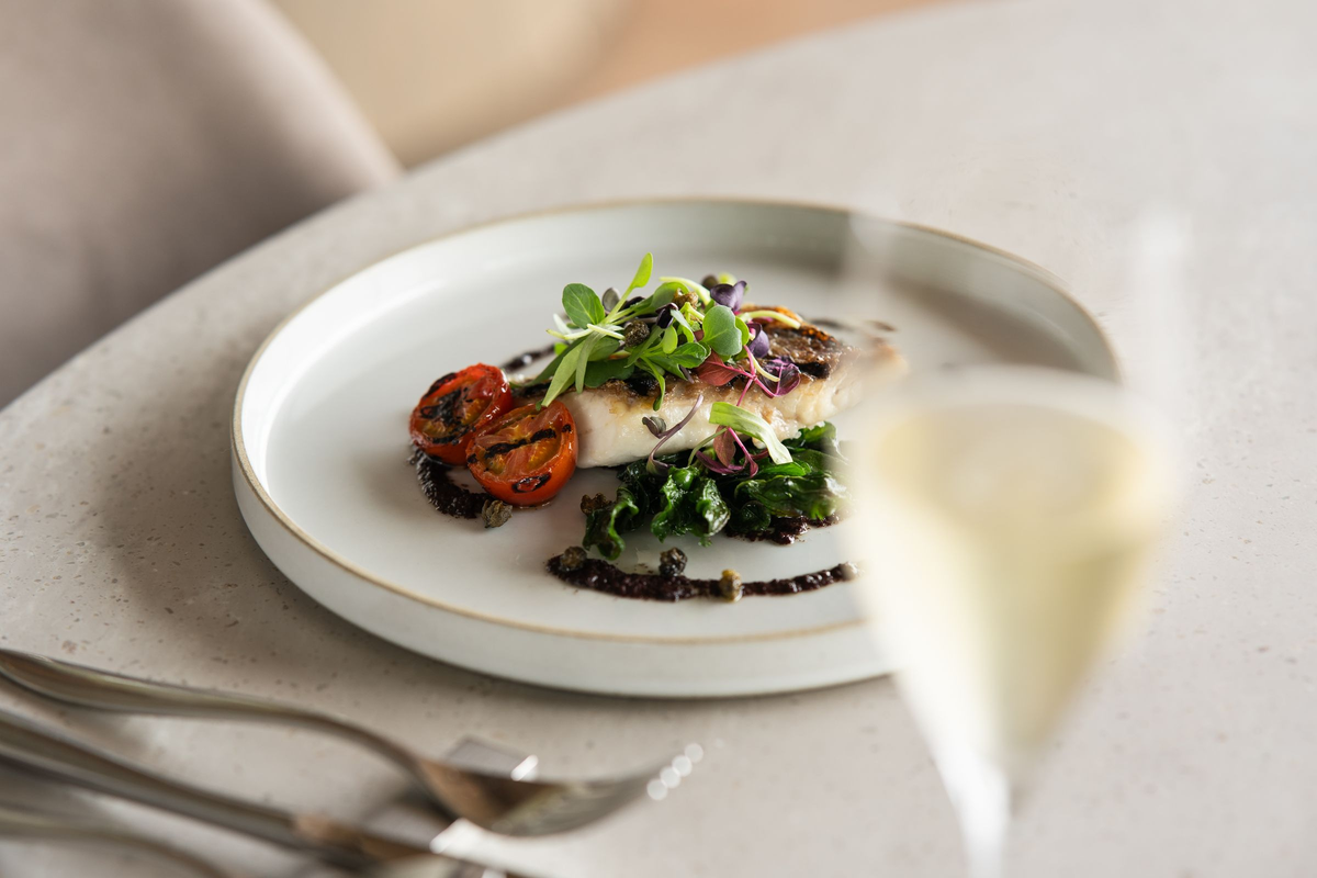 Peixe com vegetais e folhas em prato branco, acompanhado de taça de vinho branco em primeiro plano.