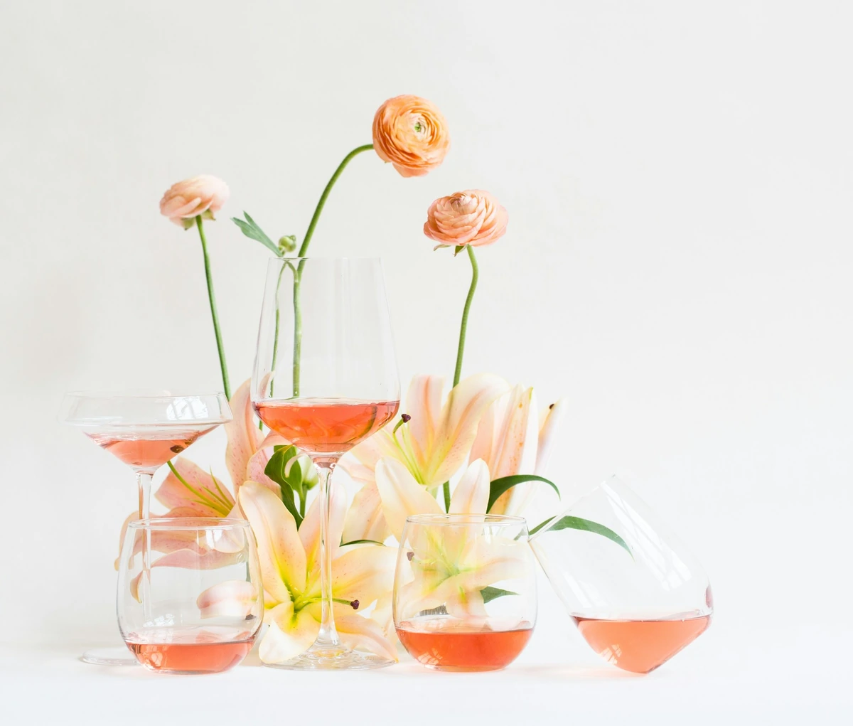 Taças e copos com bebida rosé entre flores claras e pêssego.