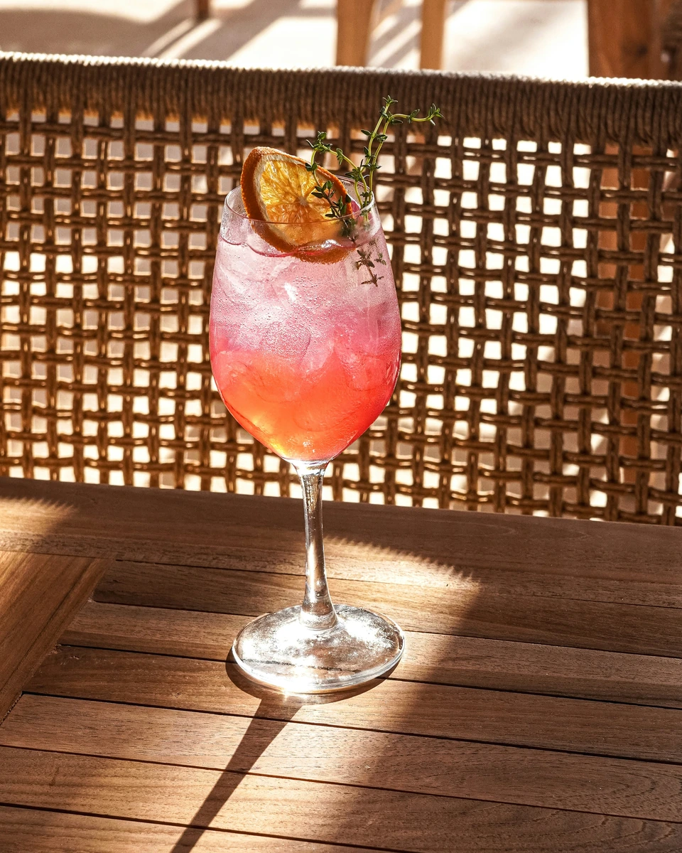Coquetel rosado com gelo, rodela cítrica e erva aromática sobre mesa de madeira.