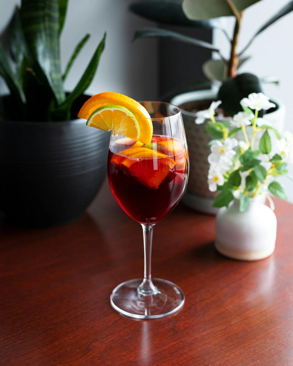 Taça de sangria rubi com fatias de laranja e limão sobre mesa de madeira.