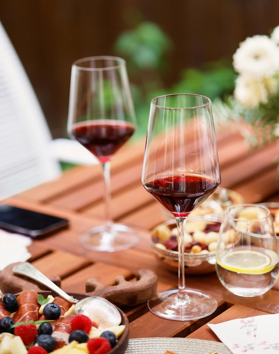 Taças de vinho tinto com tábua de frios, queijo, frutas e petiscos em mesa externa.