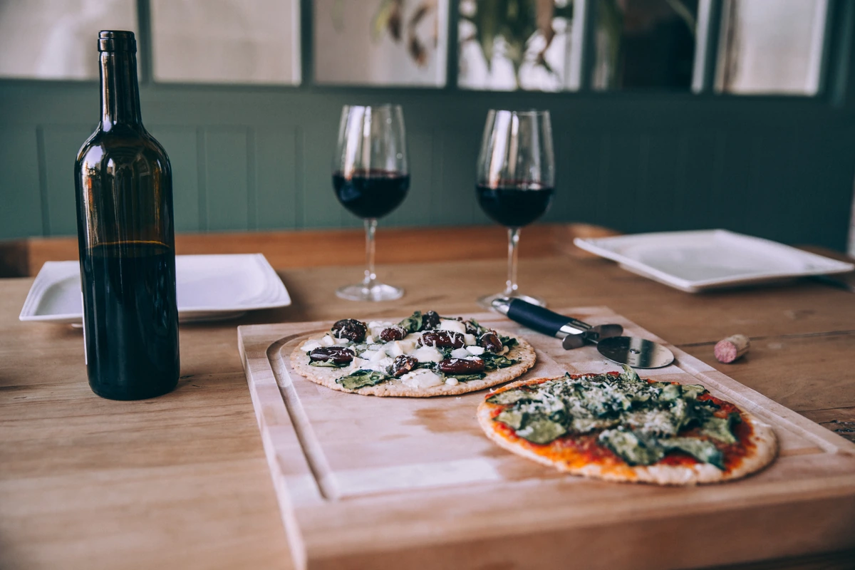 Pizzas artesanais sobre tábua de madeira com duas taças preenchidas de vinho tinto ao fundo.