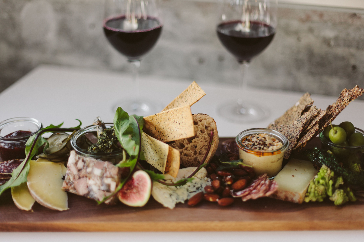 Tábua de queijos e charcutaria com pães, crackers e figo, acompanhada de duas taças de vinho tinto.