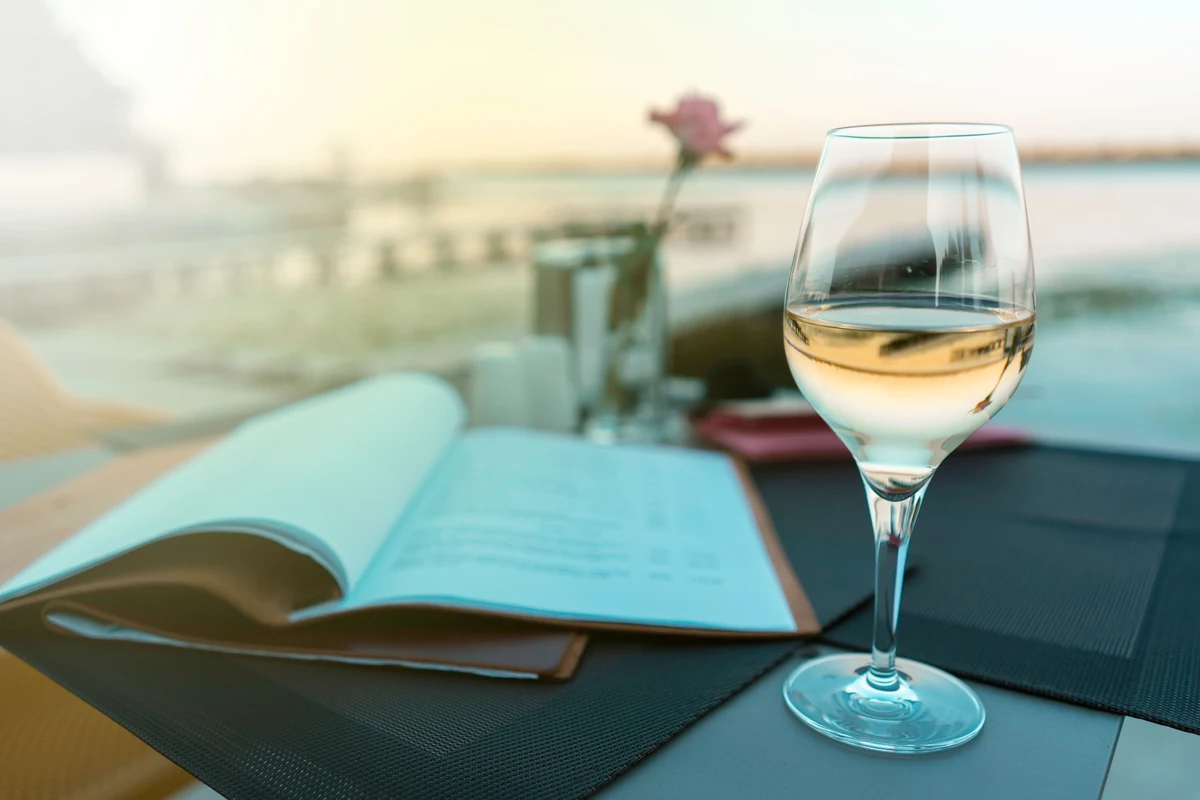 Taça de vinho branco numa mesa externa com cardápio e água ao fundo.