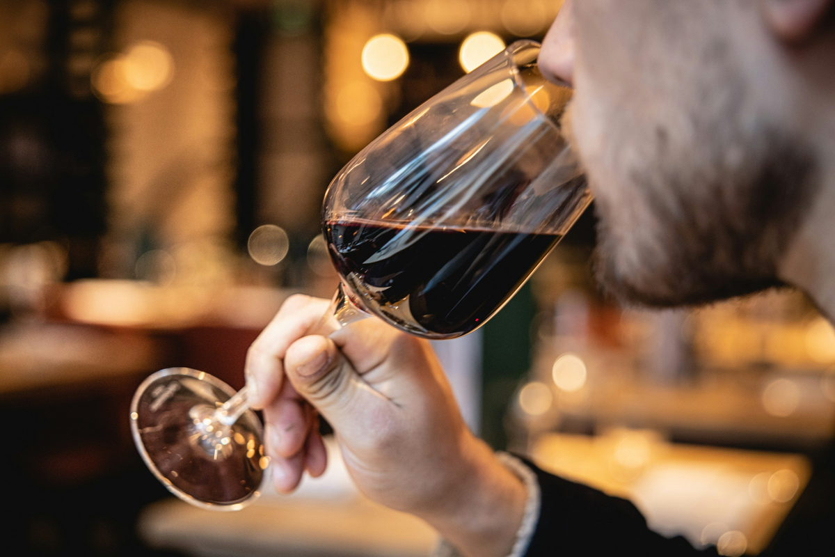 Pessoa adulta provando vinho tinto em uma taça, com iluminação quente ao fundo.
