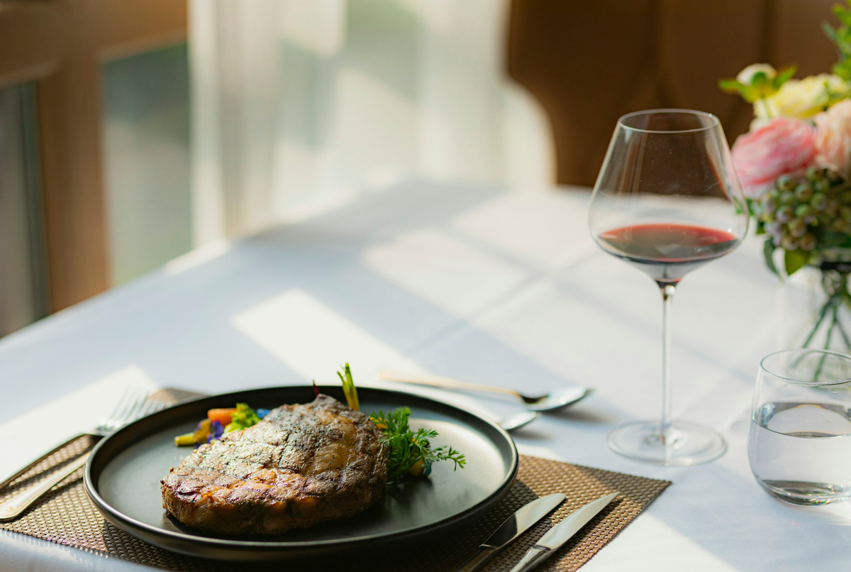 Carne grelhada com guarnição em prato escuro, ao lado de taça de tinto junto à janela.