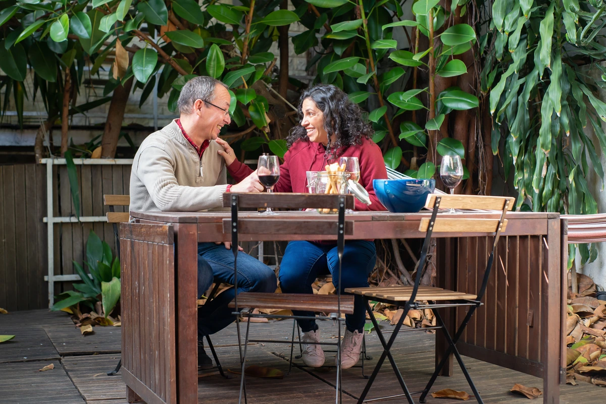Duas pessoas adultas conversam à mesa ao ar livre, com taças preenchidas de vinho tinto.