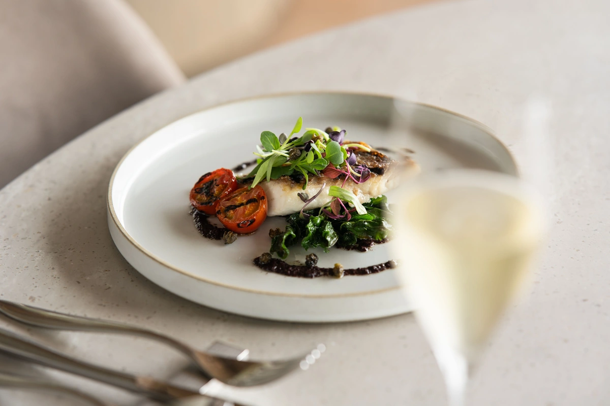 Filé de peixe com legumes no prato e taça preenchida de vinho branco desfocada em primeiro plano