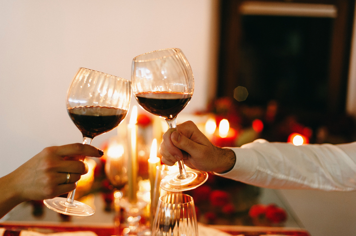 Mãos brindando com taças de vinho tinto diante de velas e flores vermelhas desfocadas.