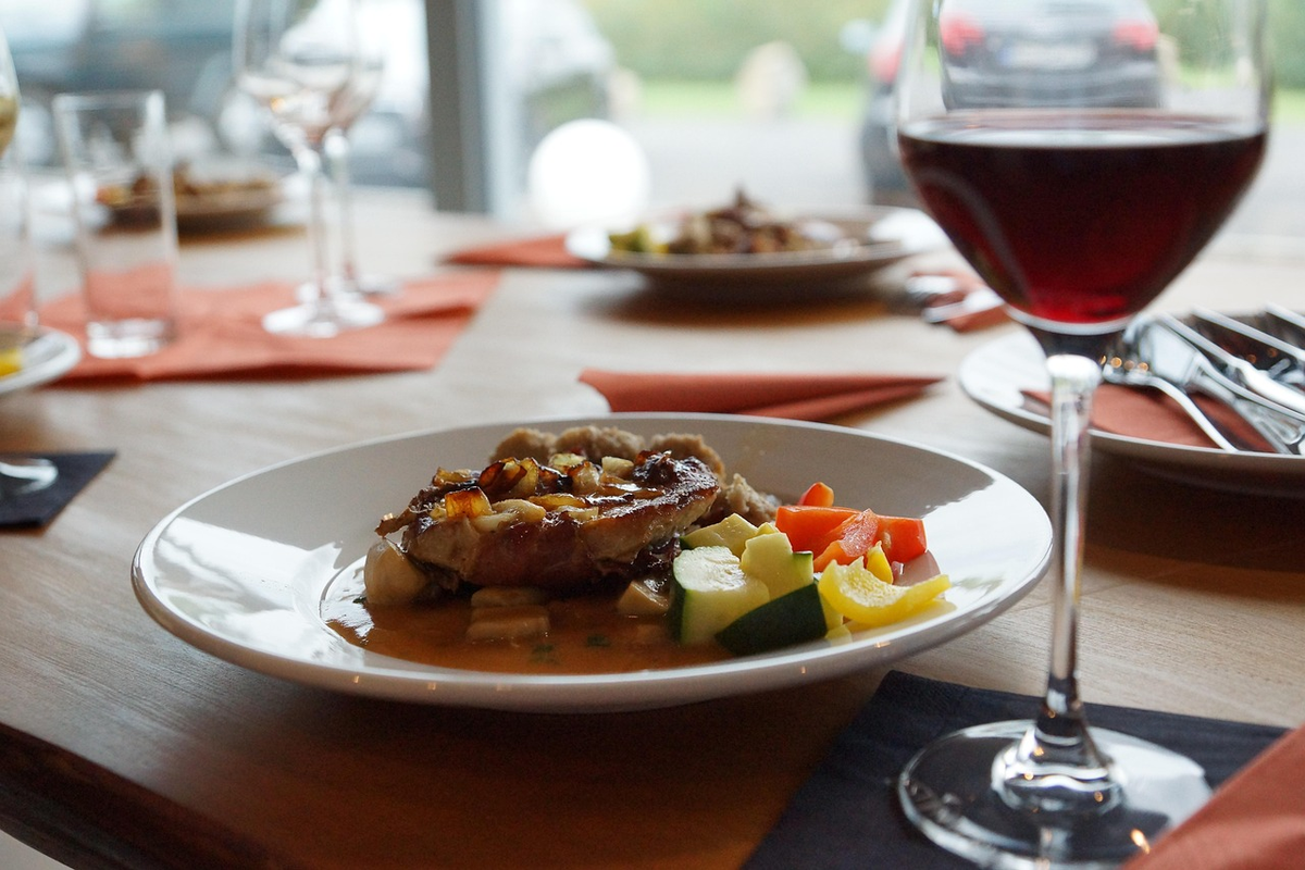 Carne dourada com molho, legumes e acompanhamento, ao lado de uma taça de vinho tinto.