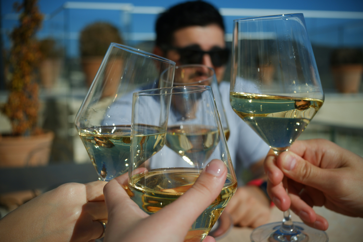 Quatro taças de vinho branco erguidas em um brinde ao ar livre, com pessoas desfocadas ao fundo.