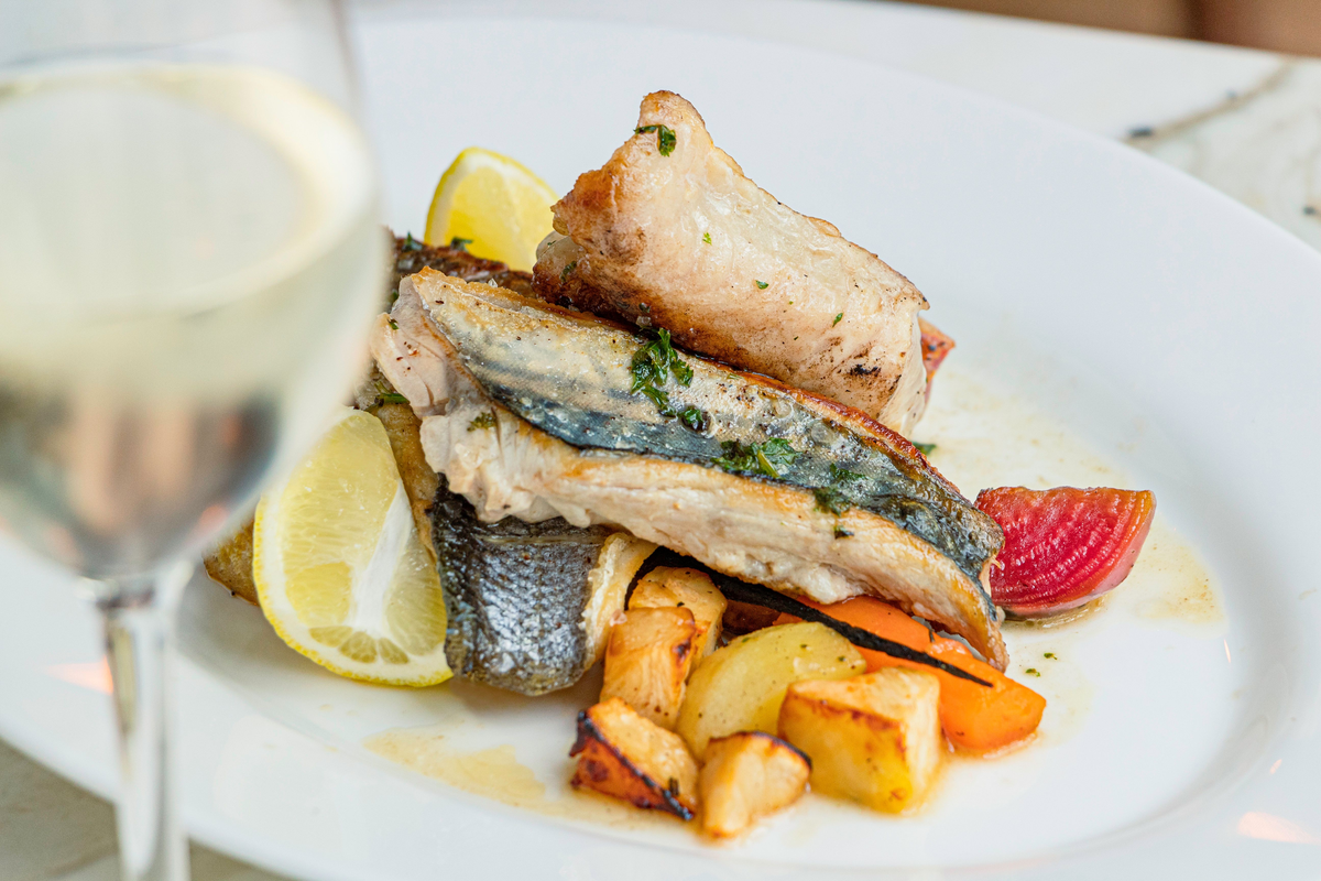 Peixe dourado com pele, batatas, legumes e limão, com uma taça de vinho branco à esquerda.