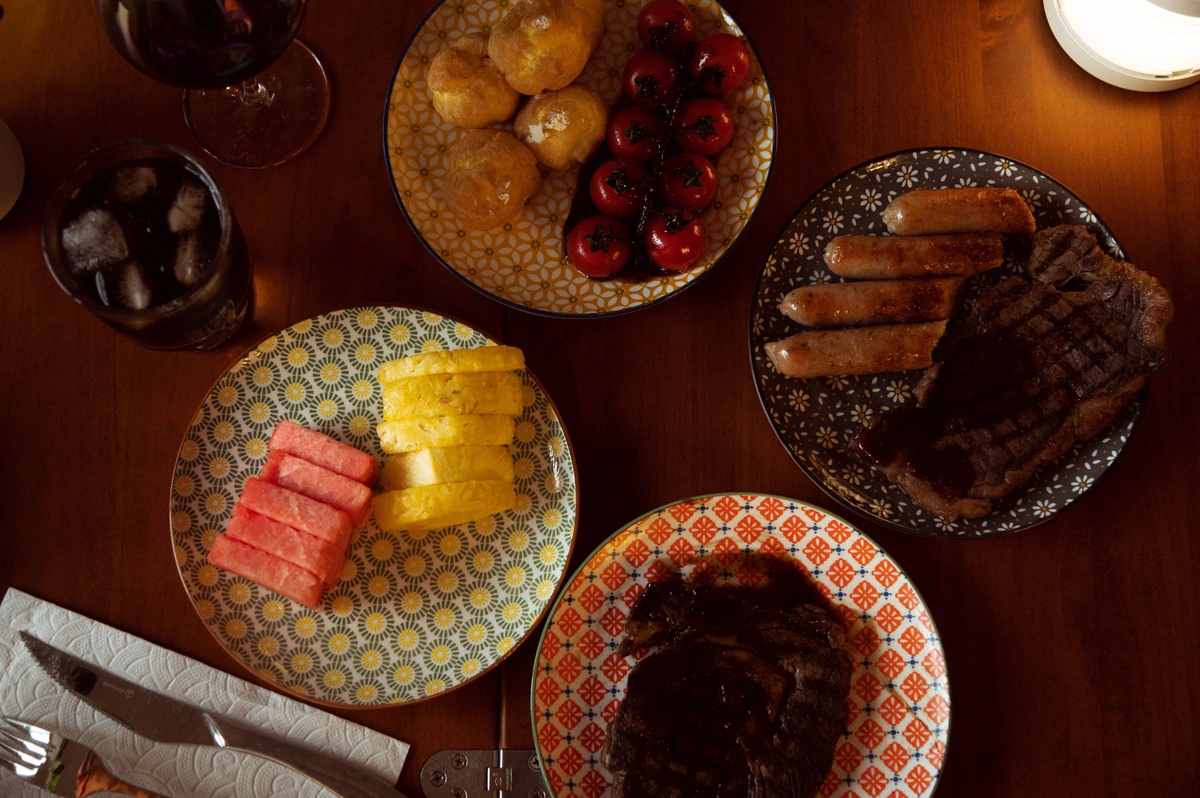 Mesa com carnes grelhadas, acompanhamentos e taça de vinho tinto parcialmente enquadrada.
