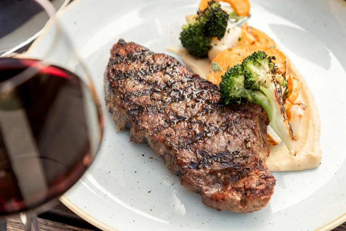 Steak bovino, legumes e purê ao lado de uma taça de vinho tinto.