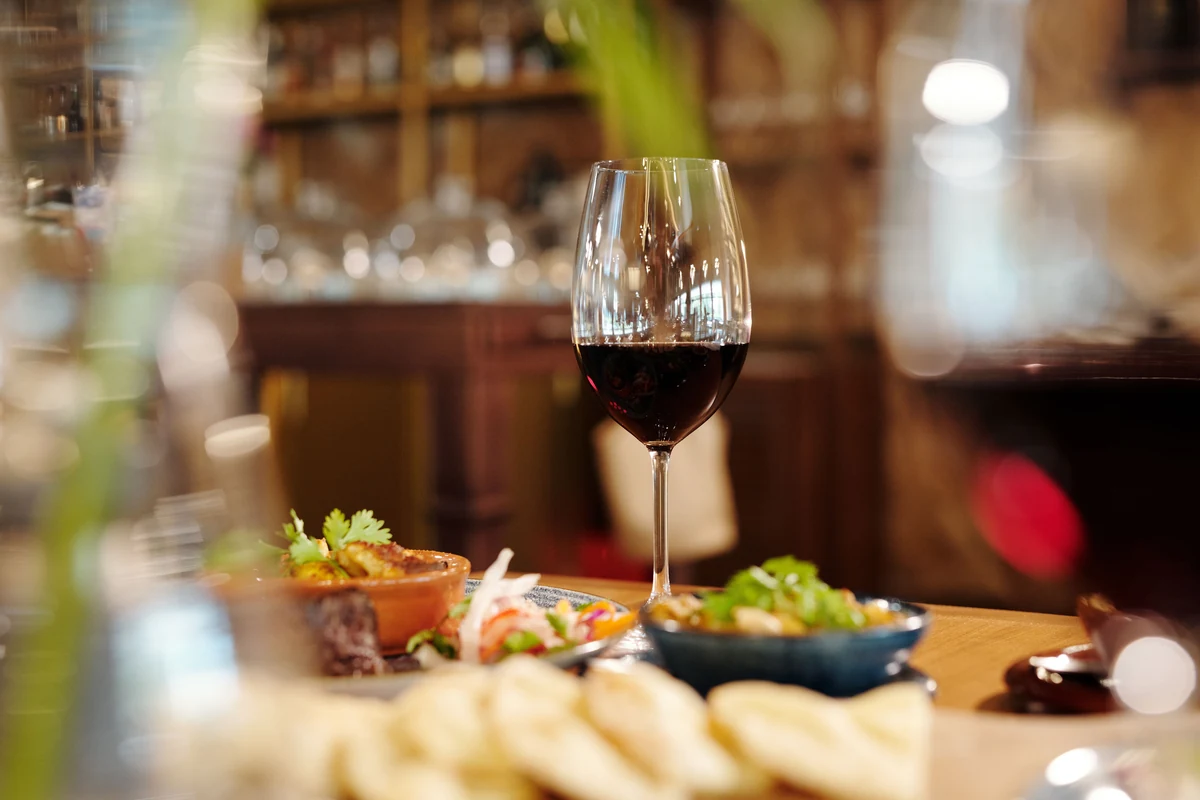 Taça de vinho tinto sobre mesa com pequenos pratos, com bar desfocado ao fundo.