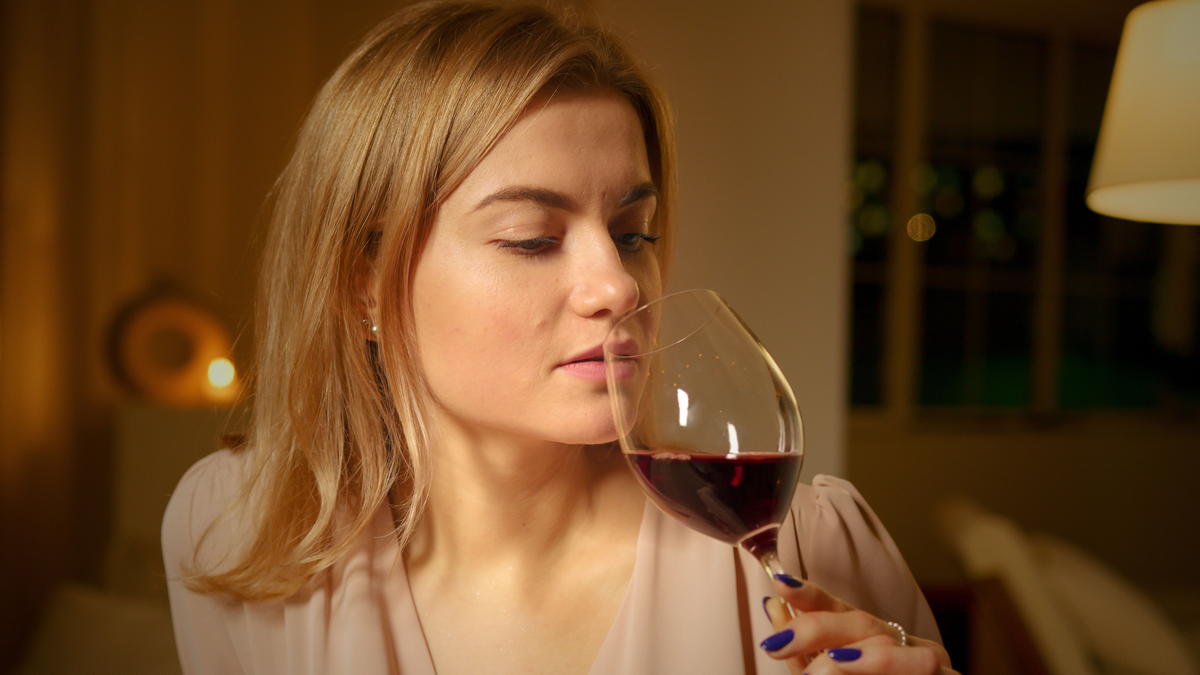 Pessoa adulta aproxima uma taça de vinho tinto do nariz em ambiente interno.
