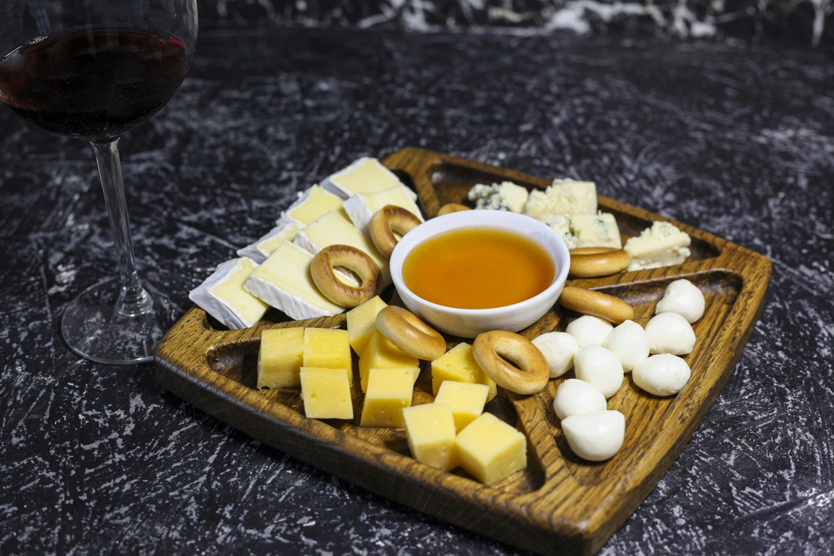 Tábua de queijos, biscoitos e mel ao lado de taça preenchida de vinho tinto.