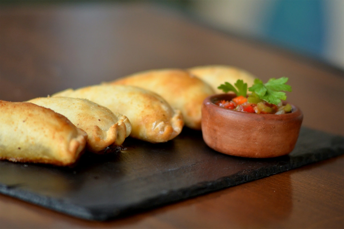 Empanadas douradas sobre ardósia, com acompanhamento em pote de barro
