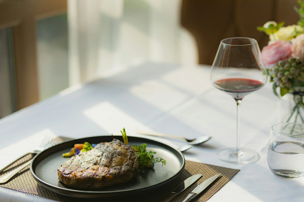 Carne bovina em prato escuro com taça de vinho tinto, linho branco e flores