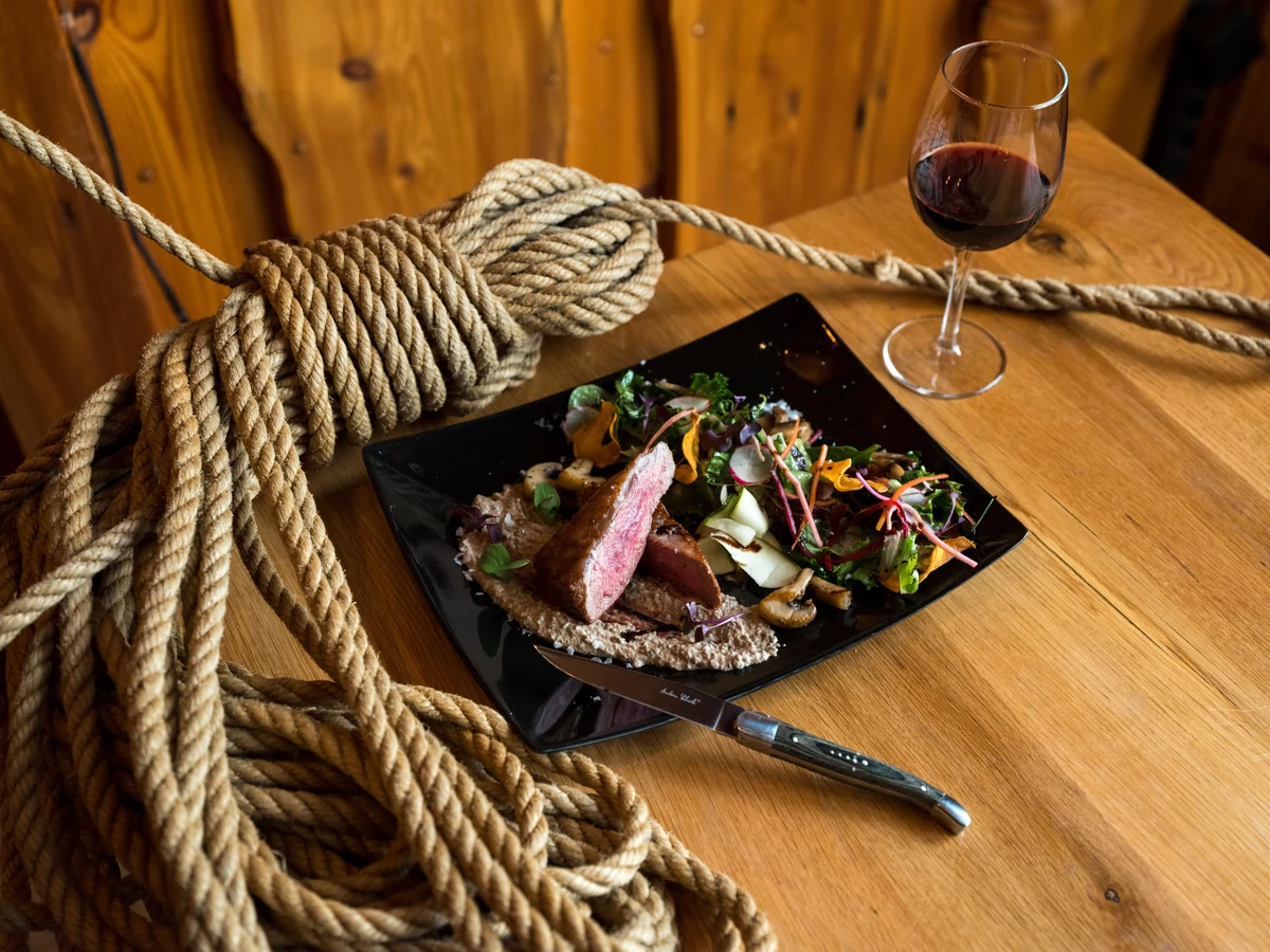 Carne fatiada com cogumelos e vegetais, junto de uma taça de vinho tinto.