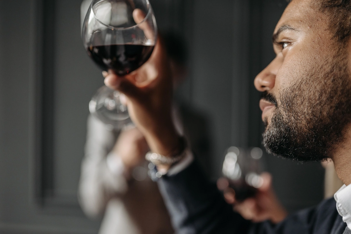 Homem observando uma taça de vinho tinto, com outra pessoa desfocada ao fundo.