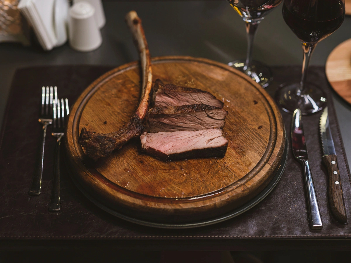 Carne bovina com osso e fatias sobre tábua, com duas taças de vinho tinto parcialmente enquadradas.