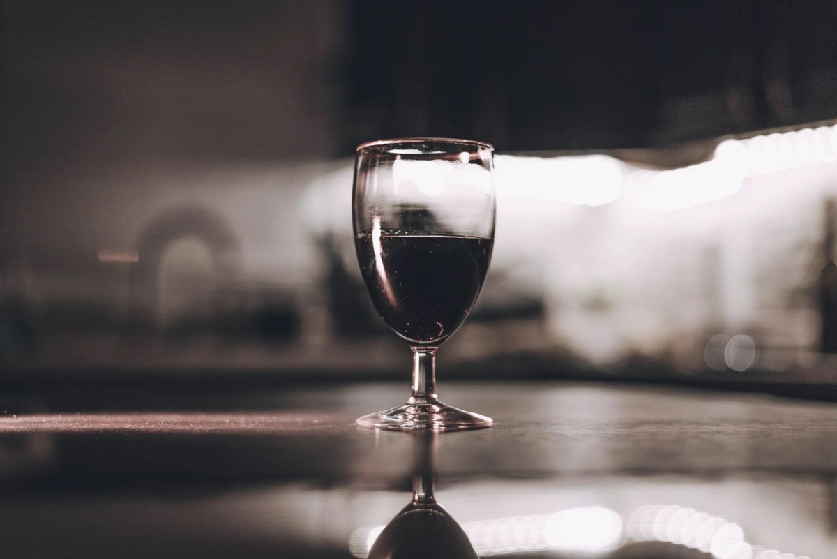 Taça com vinho tinto rubi escuro sobre mesa de madeira, com reflexos e fundo desfocado.