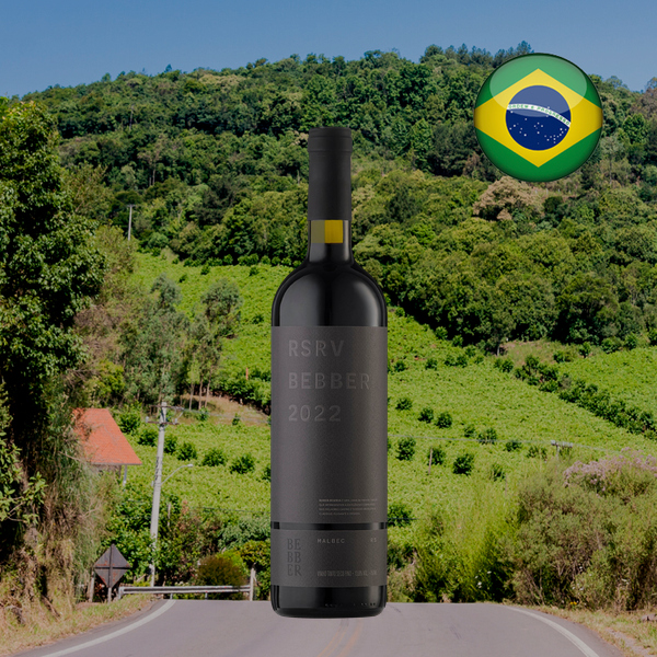 Garrafa do Família Bebber Reserva Malbec, com cápsula preta e rótulo cinza, centralizada em enquadramento quadrado diante de estrada rural, morros verdes e mata sob céu azul, com bandeira brasileira no alto à direita.