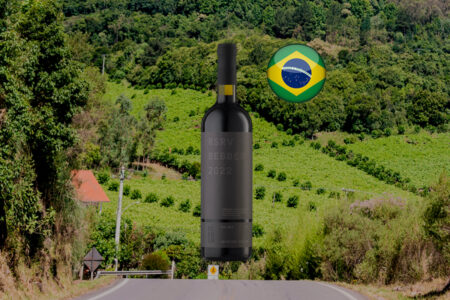 Garrafa do Família Bebber Reserva Malbec com rótulo cinza e cápsula preta, em composição horizontal diante de encosta verde com mata, telhado vermelho e estrada na parte inferior; bandeira brasileira à direita da garrafa.