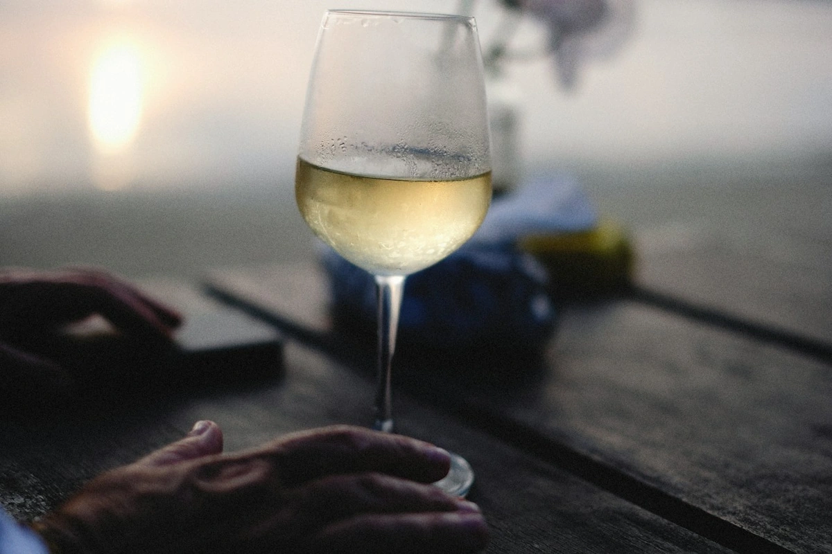 Taça com vinho branco de tom palha-dourado sobre mesa de madeira ao ar livre, com luz suave ao fundo.
