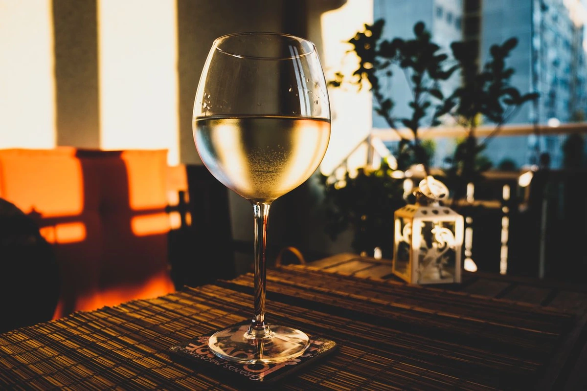 Taça com vinho branco de tom dourado-claro sobre mesa com esteira, iluminada pelo sol quente, com plantas ao fundo.