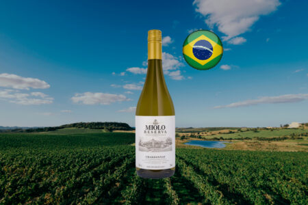 Imagem horizontal de Miolo Reserva Chardonnay, com cápsula dourada e rótulo branco, sobre vinhedos e campos com um lago ao fundo, com bandeira do Brasil.