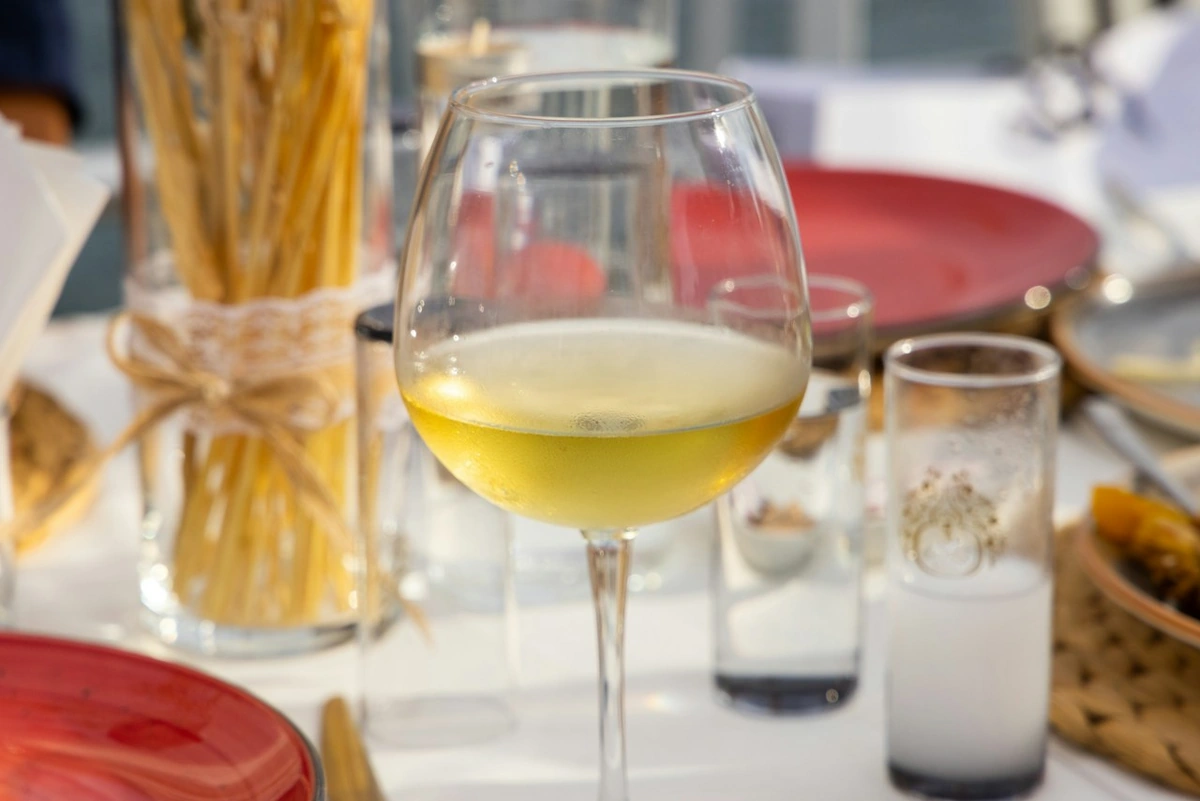 Taça preenchida com vinho branco de tom amarelo-claro em mesa posta com pratos vermelhos e recipiente de grissini.