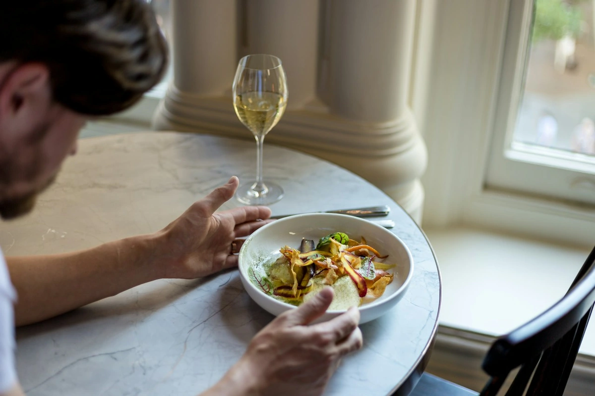 Adulto diante de um prato de massa com vegetais e molho claro, com taça preenchida com vinho branco sobre mesa junto à janela.