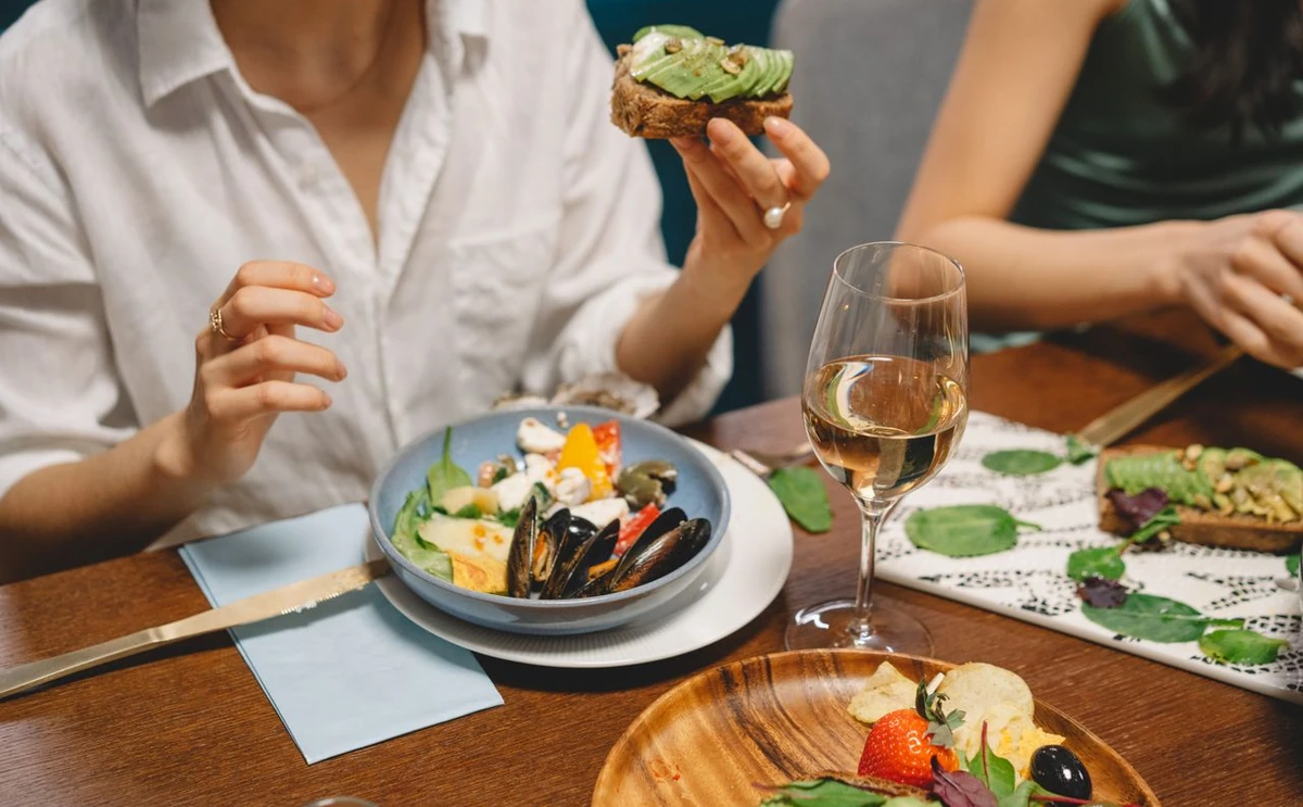 Tosta de abacate e taça com bebida clara em mesa com outros pratos, com mãos de pessoas adultas.