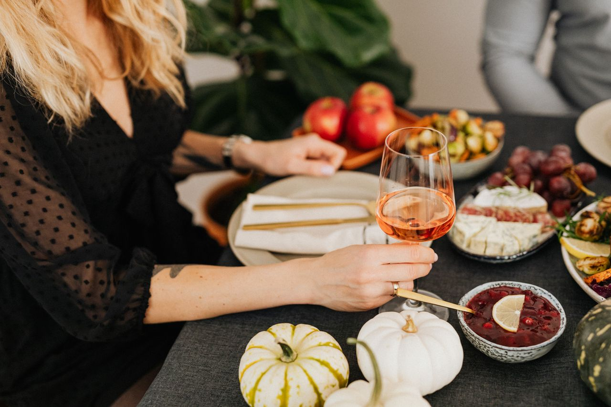 Pessoa segurando taça com vinho rosé claro à mesa com queijos, uvas, frutas e pequenas abóboras decorativas.