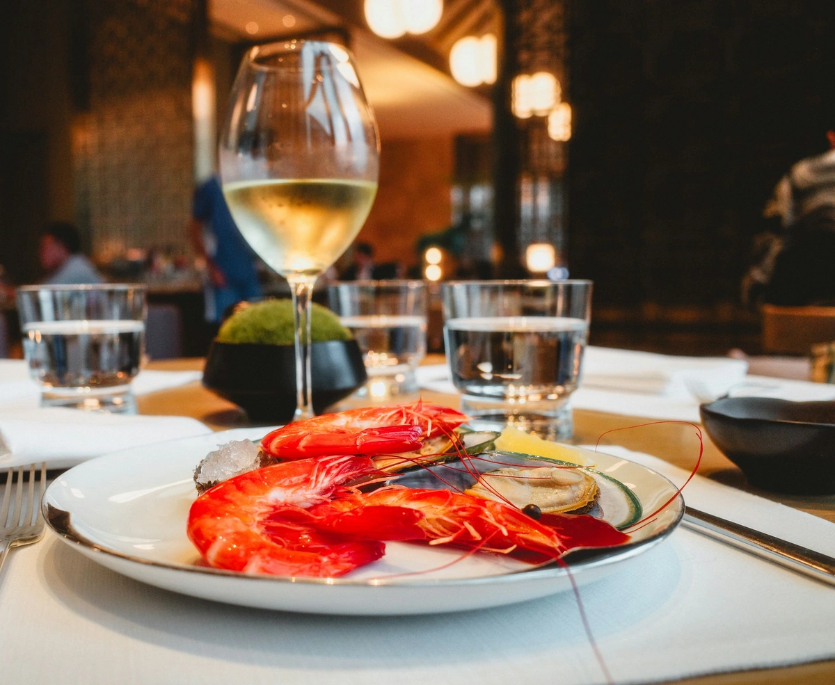 Crustáceos e moluscos com limão em prato branco, acompanhados de taça preenchida com vinho branco em mesa de restaurante.