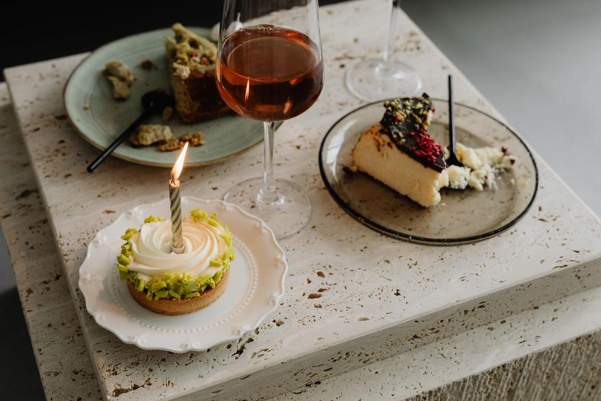 Taça com vinho rosado-acobreado ao lado de um pequeno bolo com cobertura clara e uma fatia de torta cremosa.
