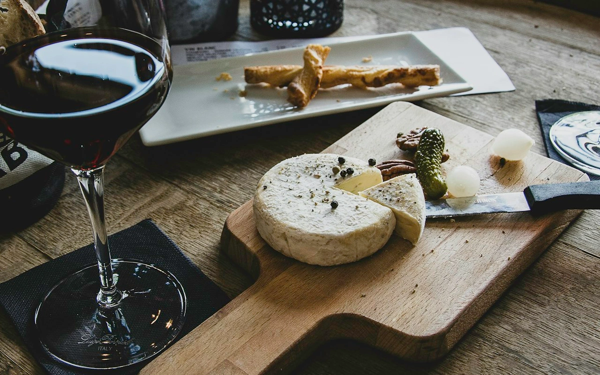 Queijo de casca branca sobre tábua com pão e conservas, ao lado de taça preenchida com vinho tinto escuro.