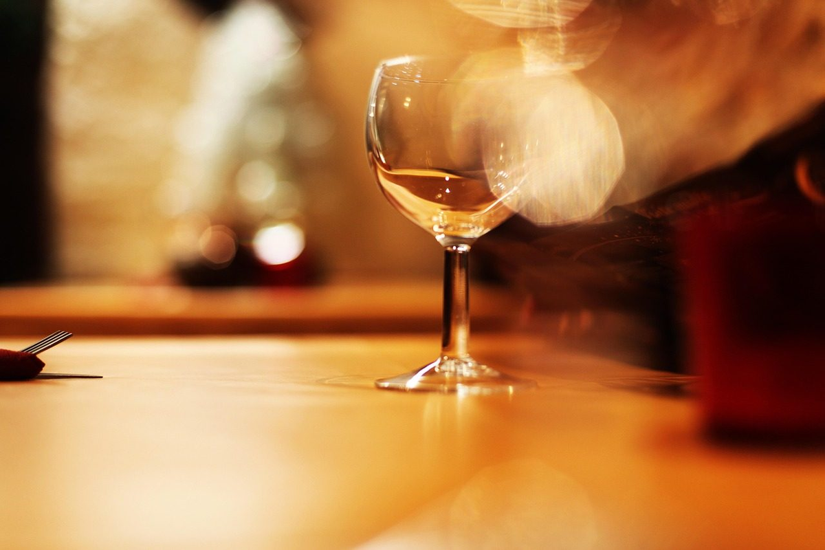 Taça com vinho branco de tom dourado sob luz quente, sobre mesa de restaurante com reflexos luminosos ao fundo.