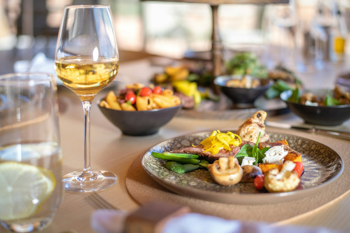 Taça com vinho branco dourado-claro ao lado de prato variado com cogumelos, folhas, legumes e outros acompanhamentos.