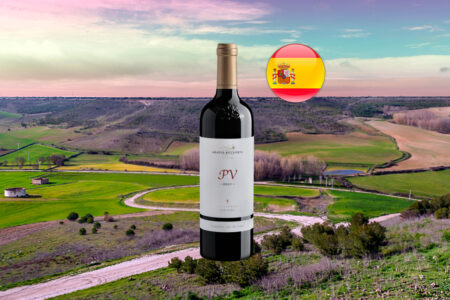 Abadía Retuerta Petit Verdot 2021 em garrafa inteira, com letras PV vermelhas no rótulo, sobre panorama horizontal de campos, caminhos curvos e céu rosado do cenário de Valladolid, com bandeira espanhola.