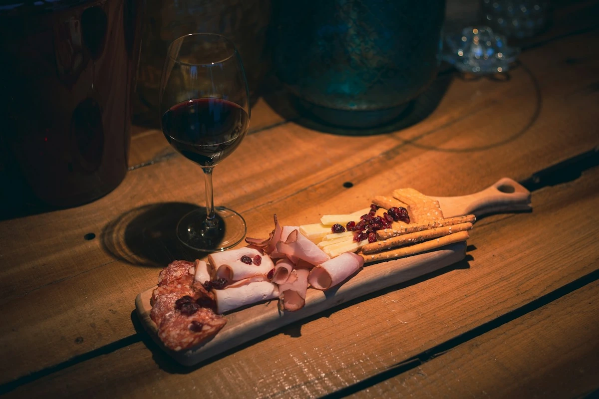 Tábua de madeira com embutidos, queijo, biscoitos e grissini, ao lado de uma taça com vinho tinto escuro.