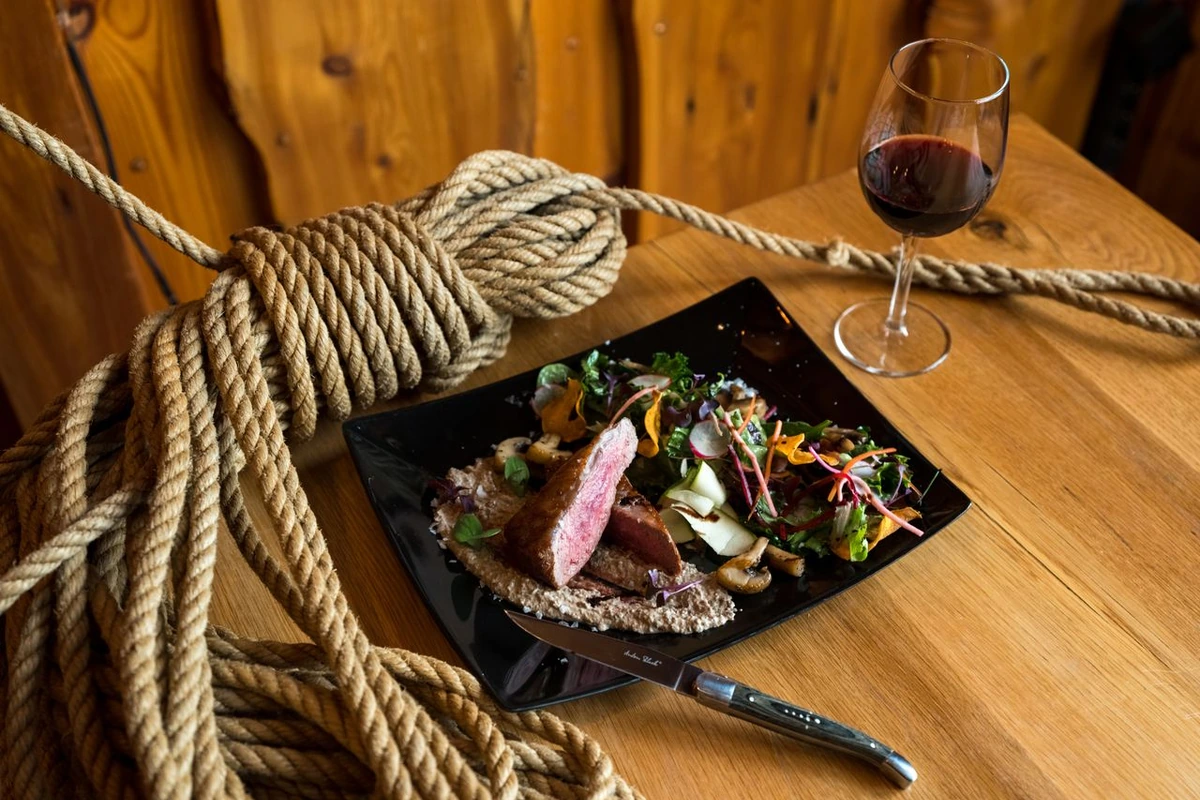 Carne bovina fatiada com molho de cogumelos e salada em prato preto, com uma taça de vinho tinto escuro ao lado sobre mesa de madeira.
