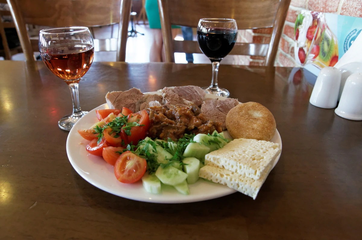 Prato com pão, tomates, pepino e acompanhamentos, com taça de vinho âmbar à esquerda e taça de tinto ao fundo.