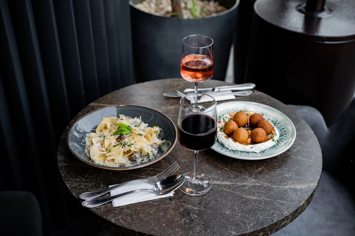 Mesa com massa, queijo e ervas, acompanhamentos e taças de vinho tinto e rosado.