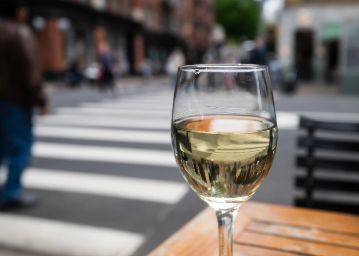 Taça de vinho branco claro em mesa externa diante de uma rua.