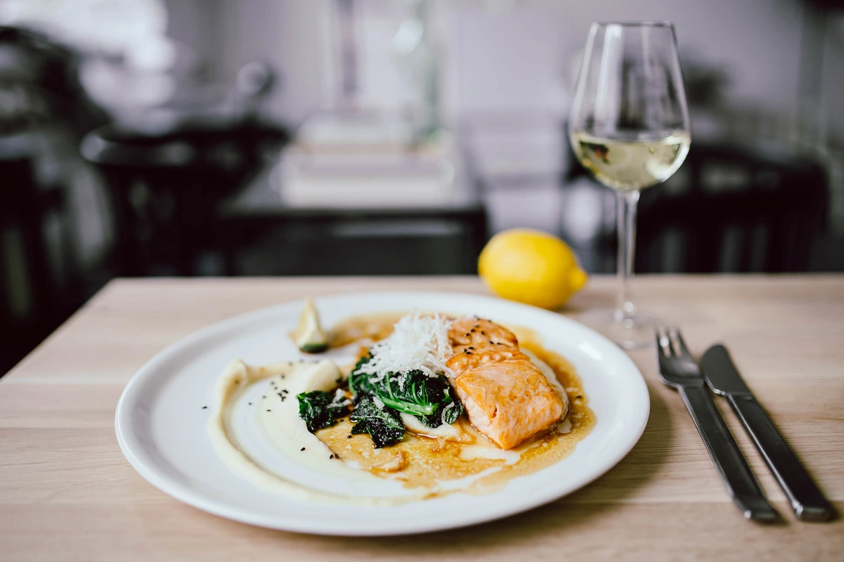 Salmão com folhas verdes e molho em prato branco, ao lado de vinho branco.